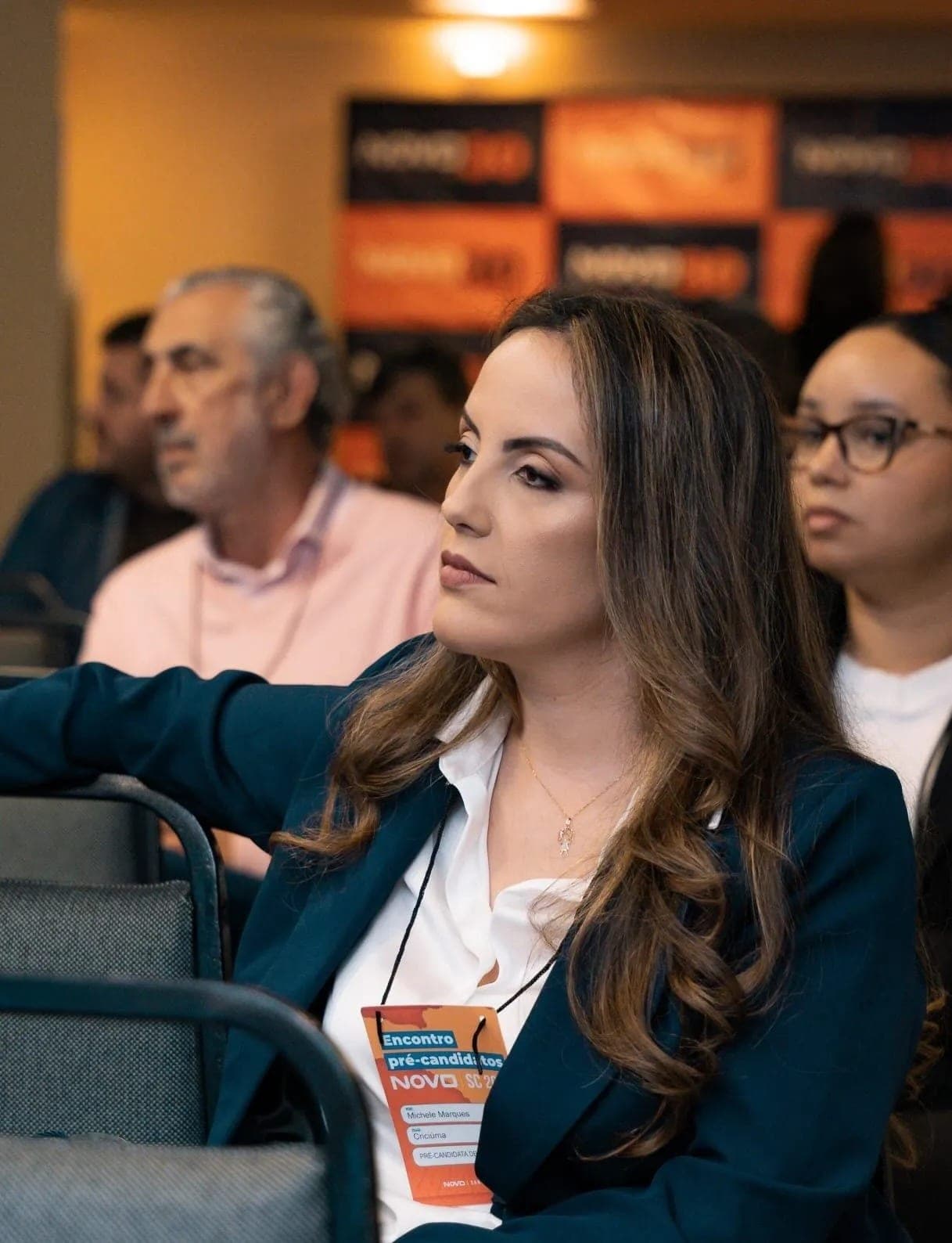 Michele Marques sentada em evento, de blazer azul e camisa branca, usando crachá do Encontro de Pré-candidatos NOVO SC