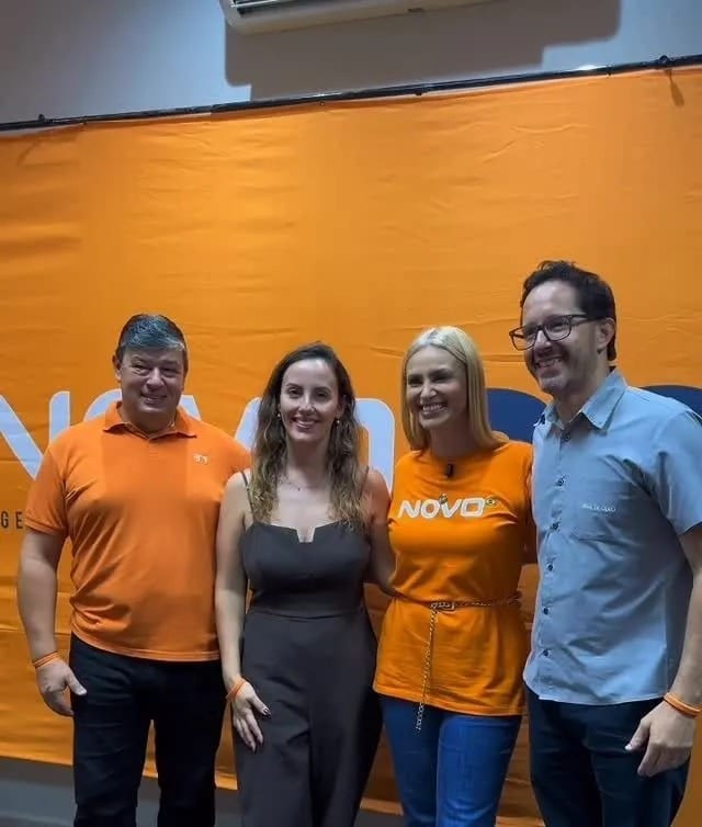 Michele Marques com três pessoas em frente a um painel laranja do NOVO