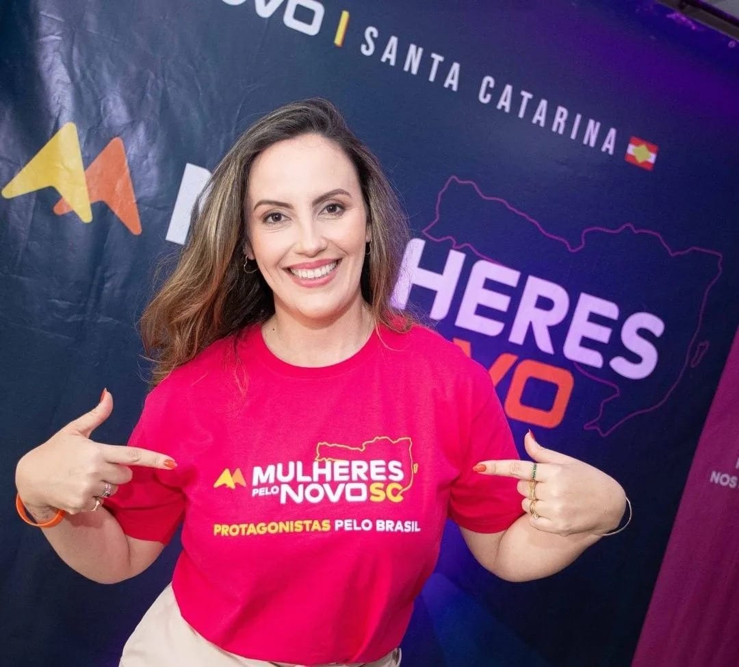 Michele Marques de camiseta Mulheres pelo NOVO SC, sorrindo em frente a um painel de Santa Catarina